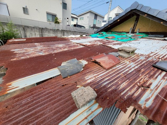 全体的に赤錆が広がったトタン屋根。瓦やブロックで応急処置がされており、雨漏りと飛散の危険がある状態。